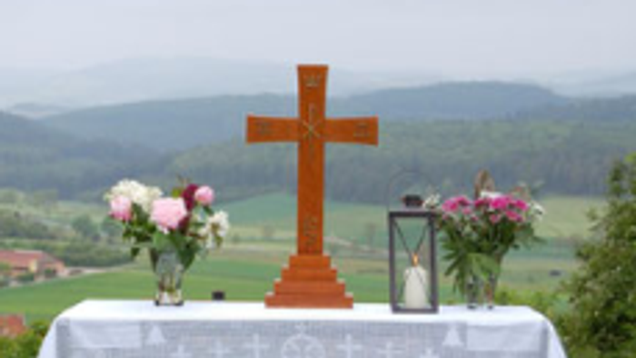 Das Foto zeigt einen aufgebauten Altar auf einer Wiese. Der Tisch trägt ein Kreuz, 2 Blumensträuße links und rechts und eine Kerze.