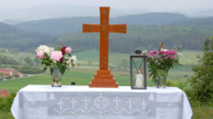 Das Foto zeigt einen aufgebauten Altar auf einer Wiese. Der Tisch trägt ein Kreuz, 2 Blumensträuße links und rechts und eine Kerze.