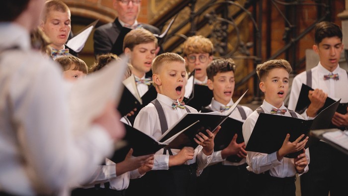 Junge männliche Chormitglieder singen leidenschaftlich in einem großen Konzertsaal mit Notenblättern.