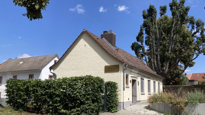 Gebäude mit Satteldach, von Hecken und Bäumen umgeben.