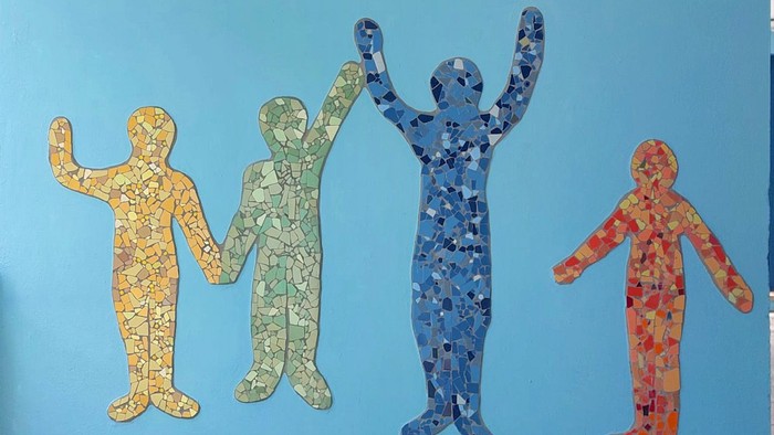 Bunte Mosaik-Wandkunst zeigt vier fröhliche Figuren, die sich an den Händen halten, sowie kleinere Herz-/Blumenfliesen darunter.