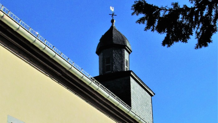 Ein gelbes Gebäude mit vergitterten Fenstern und einem dunklen Kirchturm, der von einem Kreuz gekrönt wird, unter einem klaren blauen Himmel.
