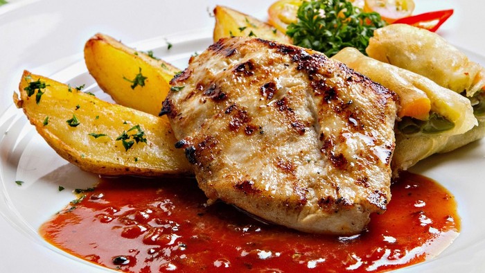 Grillet kylling med ovnstegte kartofler, indpakkede dumplings og en syrlig rød sovs på en hvid tallerken.