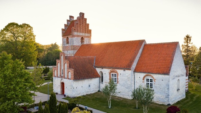 Historisk kirke med rødt tegltag og hvide stenmure i et roligt, anlagt område.
