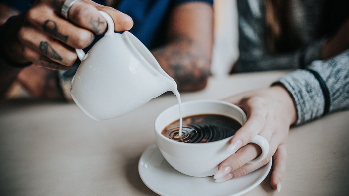 En person hælder fløde i en kop kaffe, som en anden person holder ved et bord.