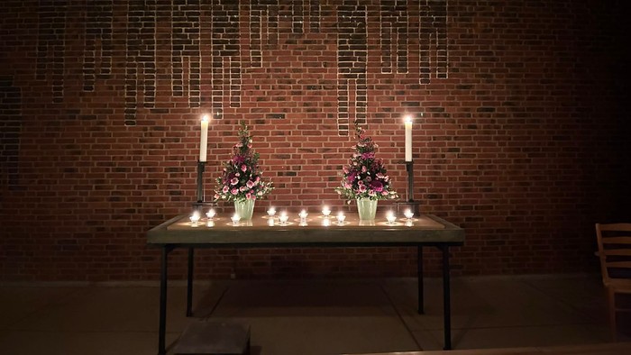 Mindesalter med tændte lys og blomsterarrangementer foran en murstensvæg med mindetavler.