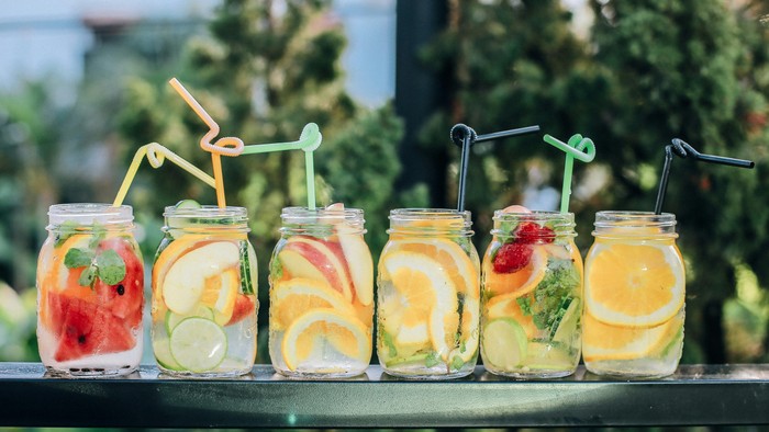 Sechs Gläser mit fruchtinfundiertem Wasser und Strohhalmen auf einem Regal.