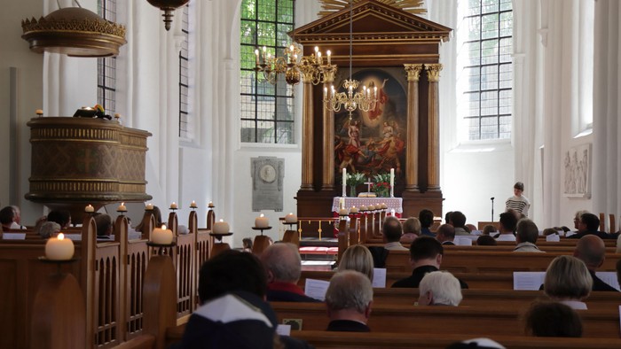 In der Kirche sitzen Menschen in Holzbänken, vorne Altar mit Kerzen und Gemälde.