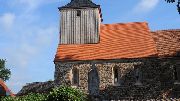 Kirchturm mit spitzem Dach und Uhr, umgeben von Steinmauer und Büschen