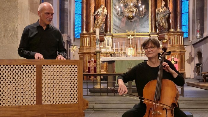 Zwei Musiker in schwarzer Kleidung musizieren in einer barocken Kirche vor einem reich verzierten Altar mit goldenen Leuchtern und blauen Fenstern. Links spielt ein Mann an einer kleinen Truhenorgel, rechts sitzt eine Frau mit einem Barockcello. Warmes Li
