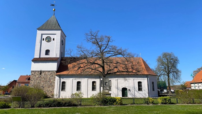 Idyllische Kirche mit hohem Glockenturm vor einem klaren blauen Himmel und üppiger grüner Umgebung.