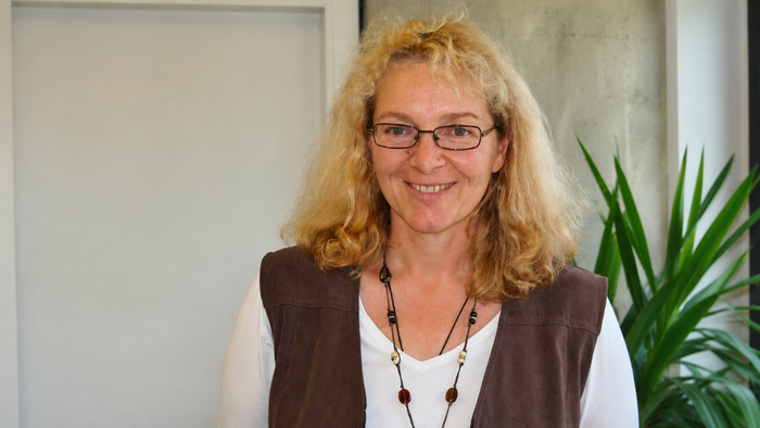 Eine fröhliche Frau mit lockigem Haar, einer Brille und einer Halskette steht drinnen in der Nähe einer Pflanze.