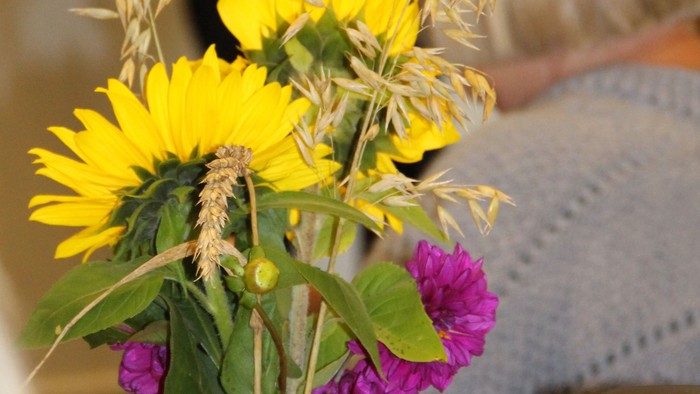 I forgrunden et bord med en buket af rødgule blomster og kornaks på et bord med kaffekande og Højskolesangbøger. 
