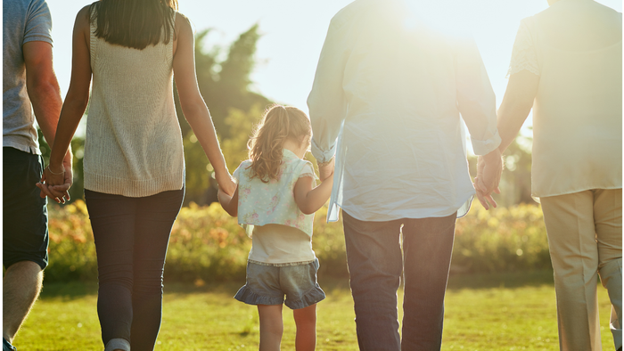 Eine Familie geht Hand in Hand durch einen sonnigen, grasbewachsenen Park und hält ein kleines Kind.