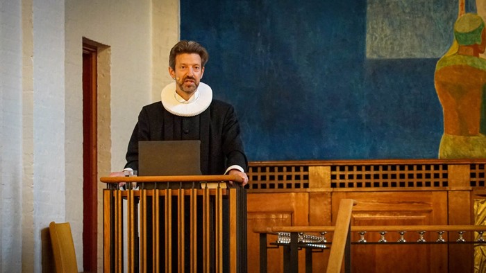 En mand i mørkt tøj og halsbånd står ved et podium og holder en laptop.