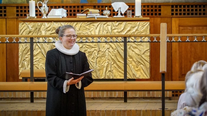En præst i sort liturgisk klæde holder en bog og står foran alteret i en kirke.
