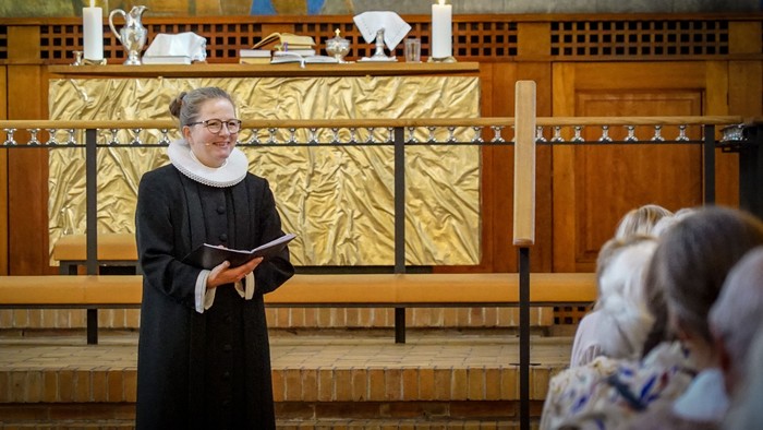 En præst i sort liturgisk klæde holder en bog og står foran alteret i en kirke.