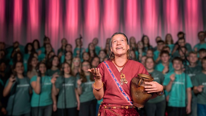 Das Bild zeigt die Hauptdarstellerin des Musicals mit einem Tonkrug in der Hand, dahinter den Chor