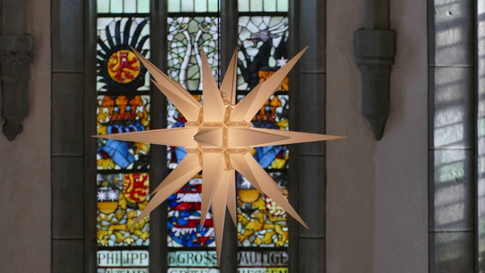 Ein großer Sternenschmuck hängt vor einem Buntglasfenster mit Text und Mustern.