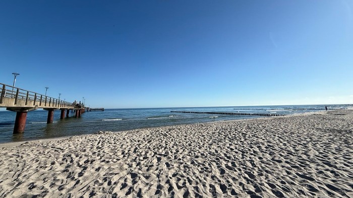 Sonnendurchfluteter Sandstrand mit einer Pier, die sich in ruhiges, klares Meerwasser unter einem strahlend blauen Himmel erstreckt.