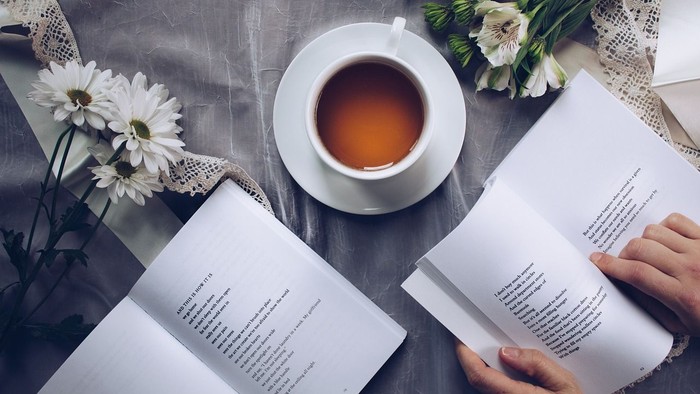 Eine Person liest in zwei Büchern, trinkt Tee und genießt Blumen auf einem Tisch.