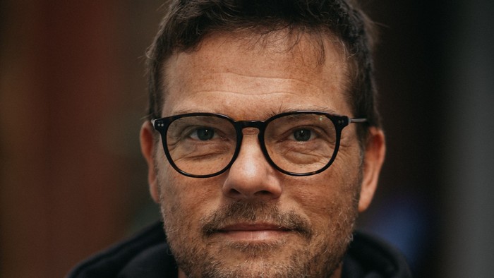 En mand med briller iført en sort hættetrøje med hvid tekst, der poser mod en mørk baggrund.