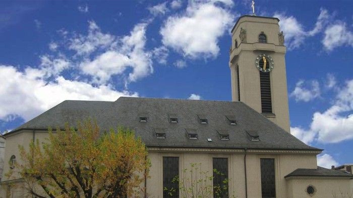 Moderne Kirche mit hohem Uhrturm vor einem strahlend blauen Himmel mit vereinzelten Wolken.