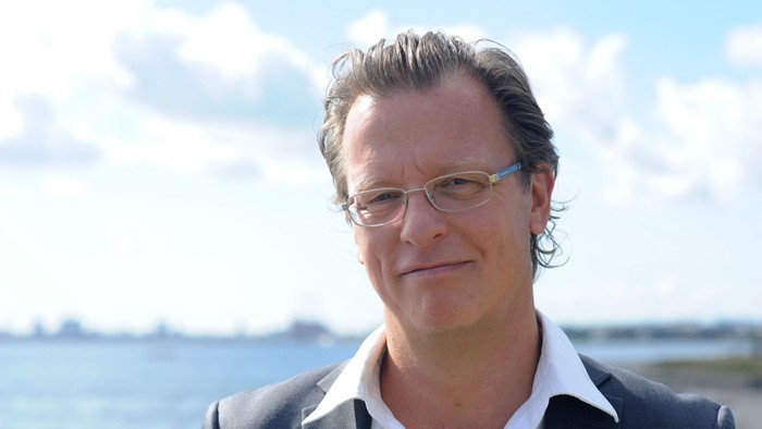 En mand med briller og jakkesæt poser foran en naturskøn havnefront med en delvist overskyet himmel.