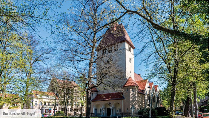 Bild zeigt eine Kirche mit hohem Turm und rotem Dach, umgeben von Bäumen und einem klaren blauen Himmel.