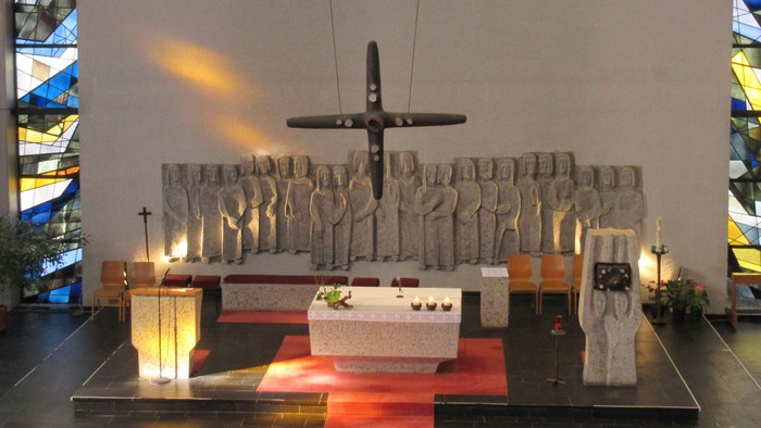 Moderner Kirchenraum der Allerheiligenkirche mit zentralem Steinaltar, einem schwebenden Bronzekreuz und einem großen Steinrelief von Heiligenfiguren im Hintergrund.