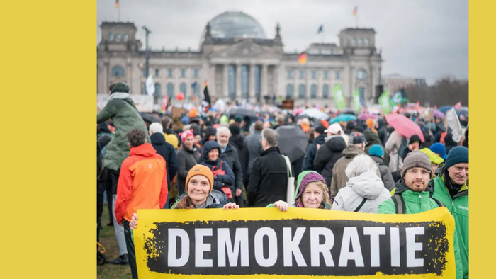 Eine große Menschenmenge versammelt sich vor einem historischen Gebäude und hält ein Schild mit der Aufschrift „Demokratie“, um sich für die Demokratie einzusetzen.