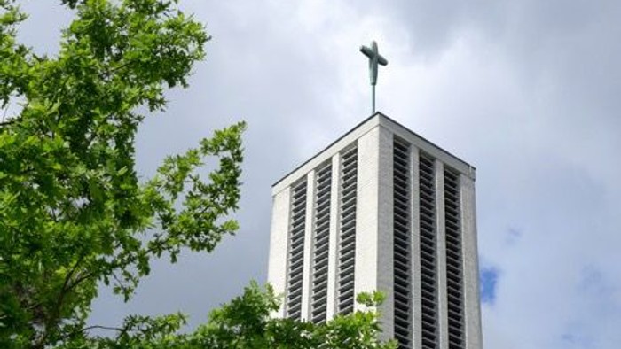 Moderner Kirchturm mit Kreuz, eingerahmt von üppigem grünem Laub unter einem bewölkten Himmel.