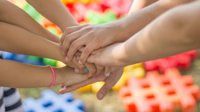 Kinderhände, die sich in einer Teamwork-Pose verbinden, mit bunten Bausteinen im Hintergrund.