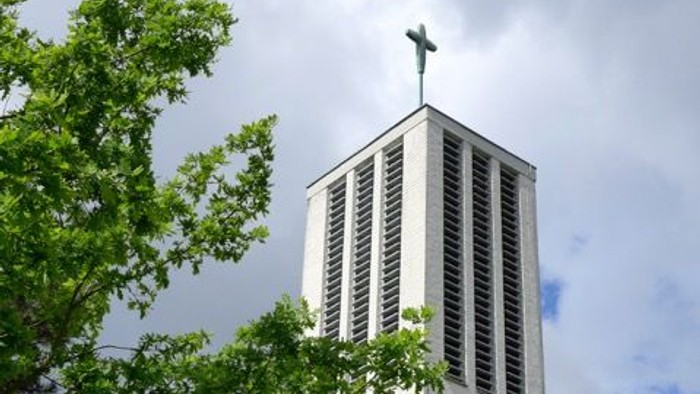 Moderner Kirchturm mit Kreuz, eingerahmt von üppigem grünem Laub unter einem bewölkten Himmel.