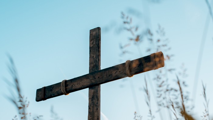 Verwittertes Holzkreuz, das hoch zwischen hohen Gräsern unter einem klaren Himmel steht.