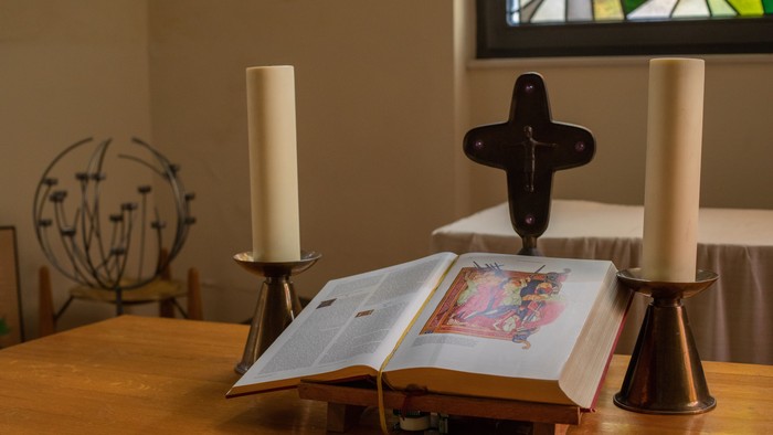 Auf einem Tisch stehen zwei Kerzen und ein aufgeschlagenes Buch mit einem Bild vor einem schwarzen Kreuz.