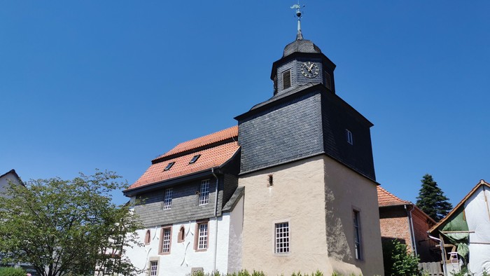 Das Bild zeigt eine Kirche mit Uhrturm und roter Ziegeldach, umgeben von grünem Gestrüpp unter klarem Himmel.