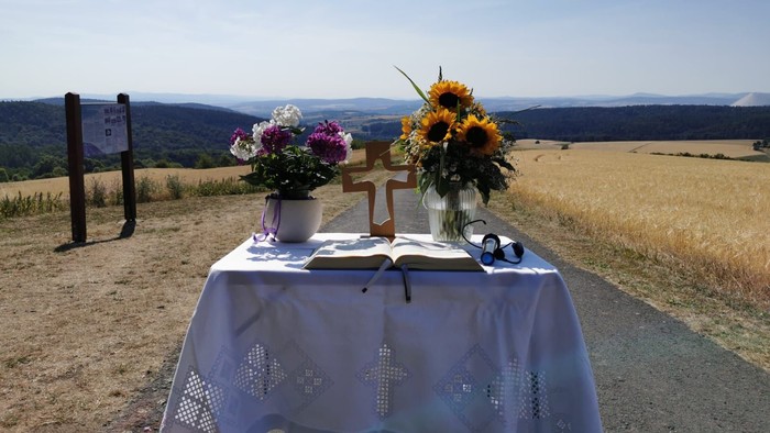 Ein Tisch mit Blumen, Kreuz und Buch steht auf einem Feld mit weitem Bergblick
