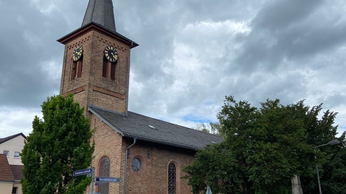 Bild der Nazarethkirche in Frankfurt Eckenheim von Südwesten her. Die Kirche hat einen spitzen Turm mit einem Kreuz . Das Dach ist mit Schiefer gedeckt. Die Wände bestehen aus gelben und roten Klinker-Steinen. In den Wänden sind große farbige Fenster mit