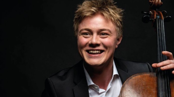 En glad musiker holder en cello og smiler til kameraet.