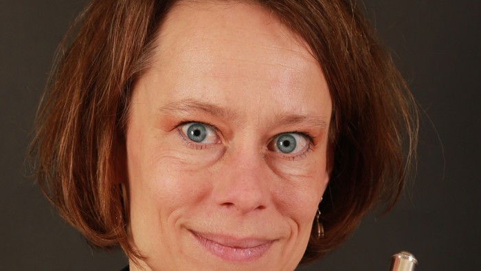 En kvinde i professionelt tøj holder og smiler med en sølvfløjte ved siden af sit ansigt.
