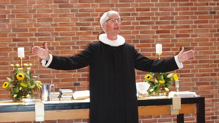 En person i religiøst tøj står med udstrakte arme og taler foran et pyntet alter.