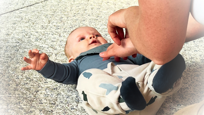 En baby i en heldragt rækker op, mens den bliver støttet af en voksens hænder på et struktureret gulv.