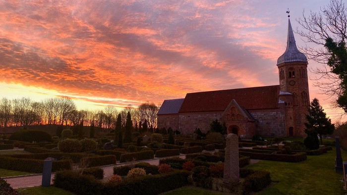 En kirke med tårn og klokke i solnedgang med rød og orange himmel, omgivet af en gravplads og grønne planter.