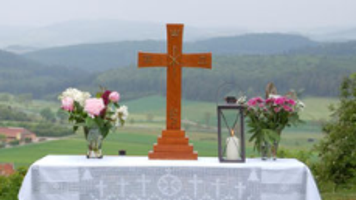 Das Foto zeigt einen aufgebauten Altar auf einer Wiese. Der Tisch trägt ein Kreuz, 2 Blumensträuße links und rechts und eine Kerze.