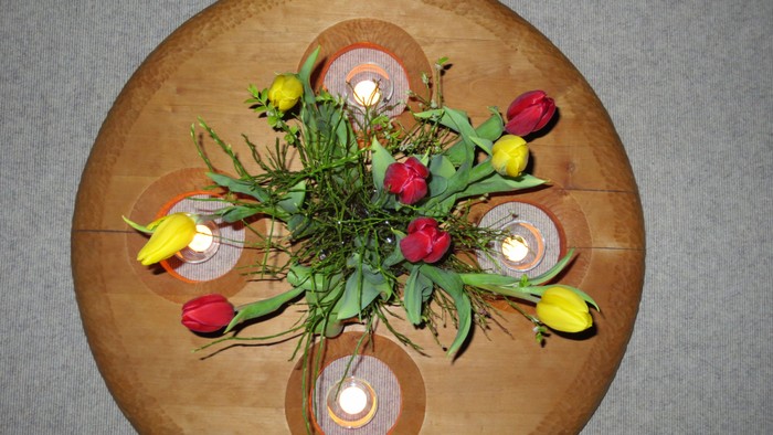 Eine rustikale Holzplatte trägt eine charmante Blumenanordnung mit Kerzen und Grünzeug in runden Haltern.
