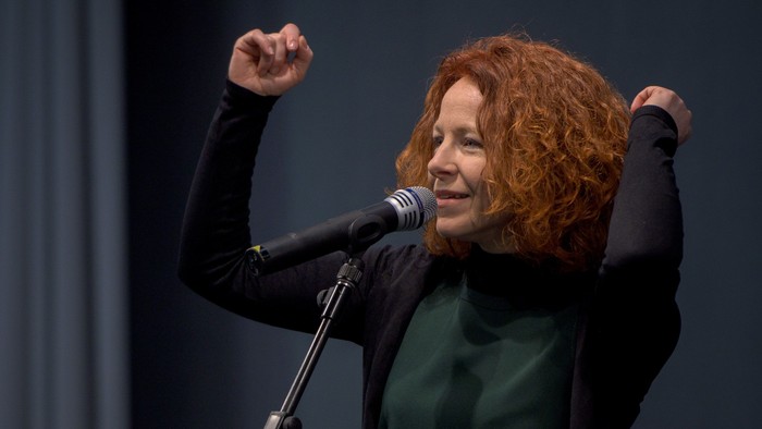 Eine Frau mit lockigem rotem Haar singt leidenschaftlich in ein Mikrofon, die Fäuste erhoben.