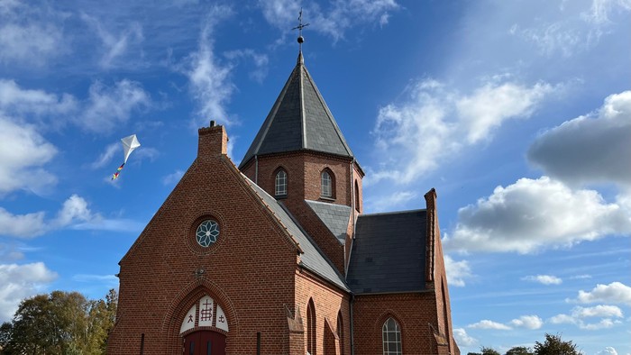 En rød murstenskirke med spids tårn og klokke under et blødt blå himmel.
