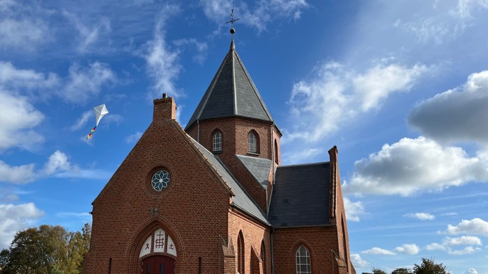En rød murstenskirke med spids tårn og klokke under et blødt blå himmel.