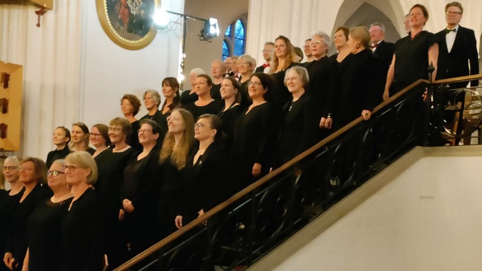 Et kor optræder sammen på en trappeopbygget balkon i en kirke, ledet af en dirigent.